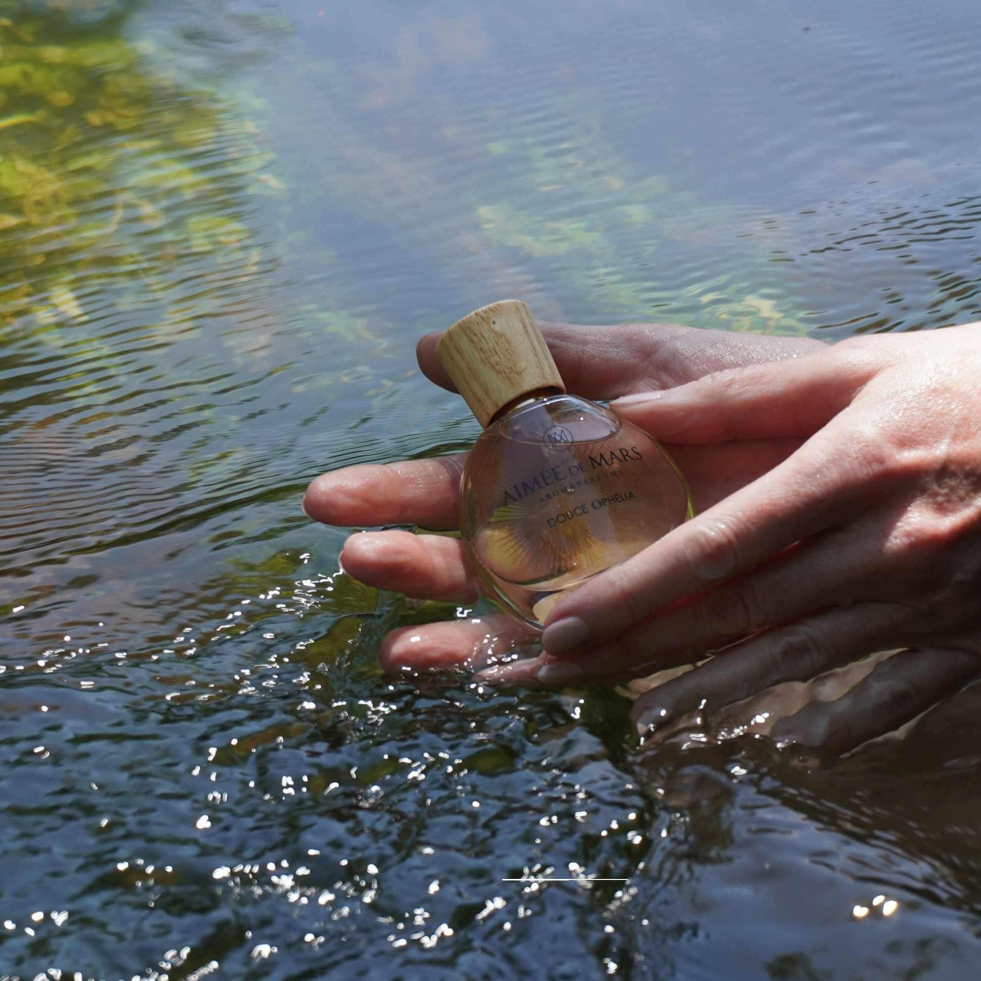 Hände halten eine Glasflasche im Wasser. Holzverschluss sichtbar. Aufschrift: Aimée de Mars, Douce Ophélia.