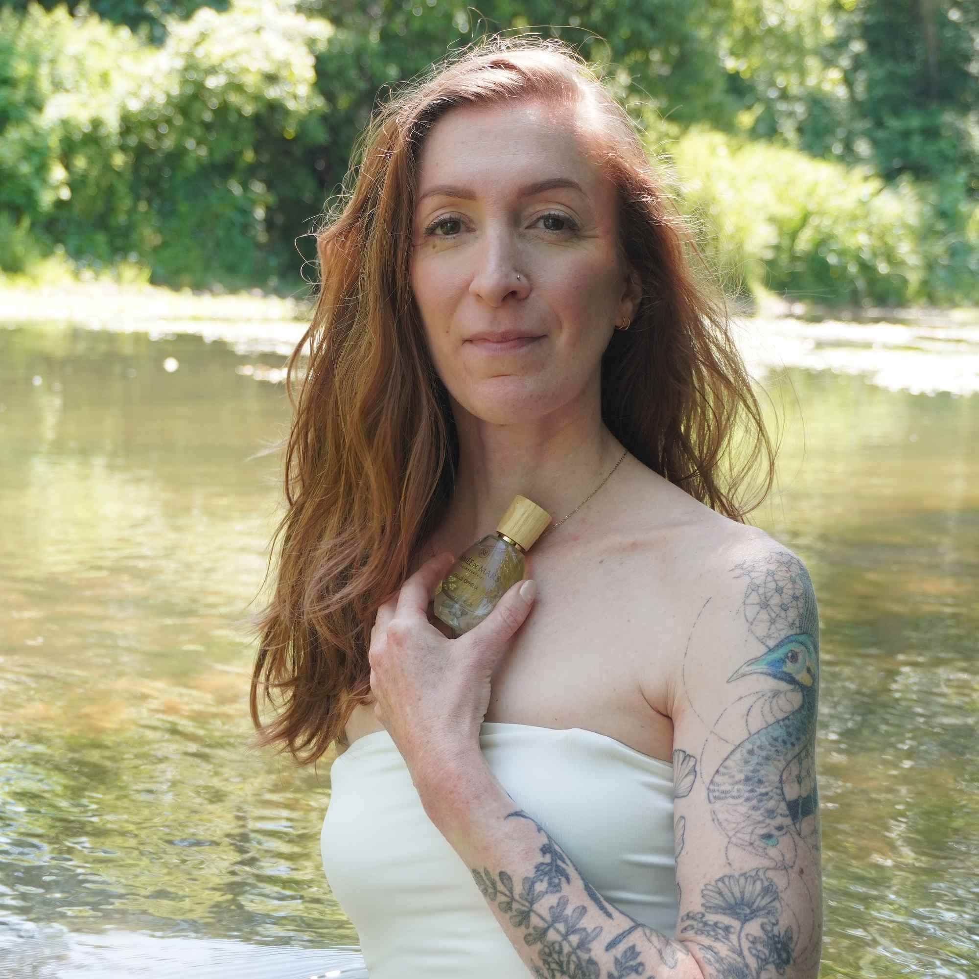 Frau hält eine Glasflasche. Holzverschluss sichtbar. Aufschrift: Aimée de Mars, Douce Ophélia. Tattoo am Arm.