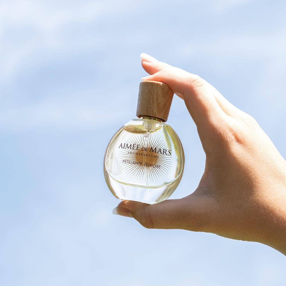 Hand hält einen runden Parfümflakon. Aufschrift: Aimée de Mars, Pétillante Aurore. Blauer Himmel im Hintergrund.