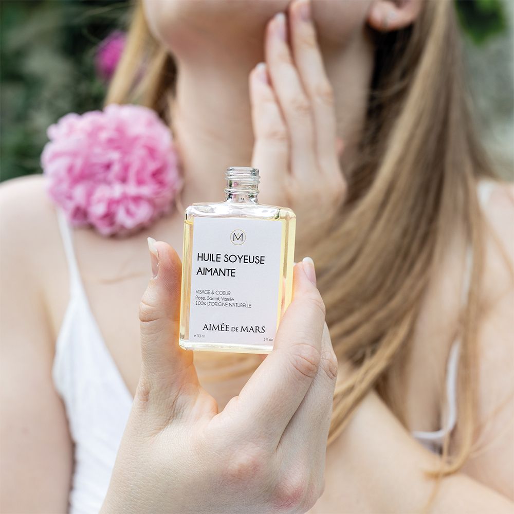 Frau hält eine Glasflasche. Aufschrift: Huile Soyeuse Aimante, Aimée de Mars. Rosa Blume im Hintergrund.