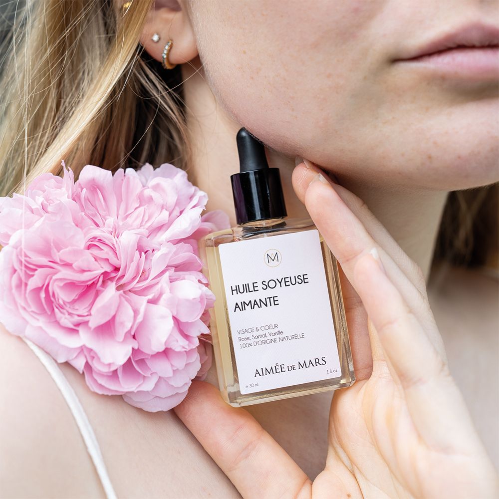 Glasflasche mit schwarzem Tropfer. Aufschrift: Huile Soyeuse Aimante, Aimée de Mars. Rosa Blume neben der Frau.