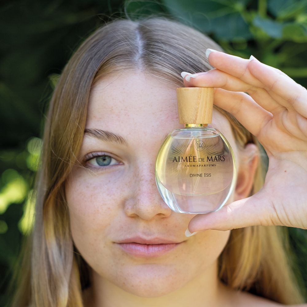 Frau hält Flakon vor Gesicht. Flakon: rund, Glas, Holzverschluss. Aufschrift: Aimée de Mars, Divine Isis. Goldfarbene Flüssigkeit.