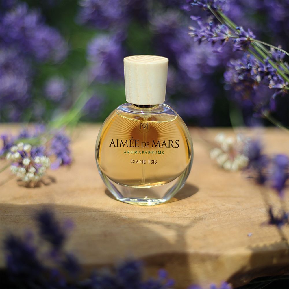 Flakon auf Holz. Flakon: rund, Glas, Holzverschluss. Aufschrift: Aimée de Mars, Divine Isis. Umgeben von Lavendelblüten.