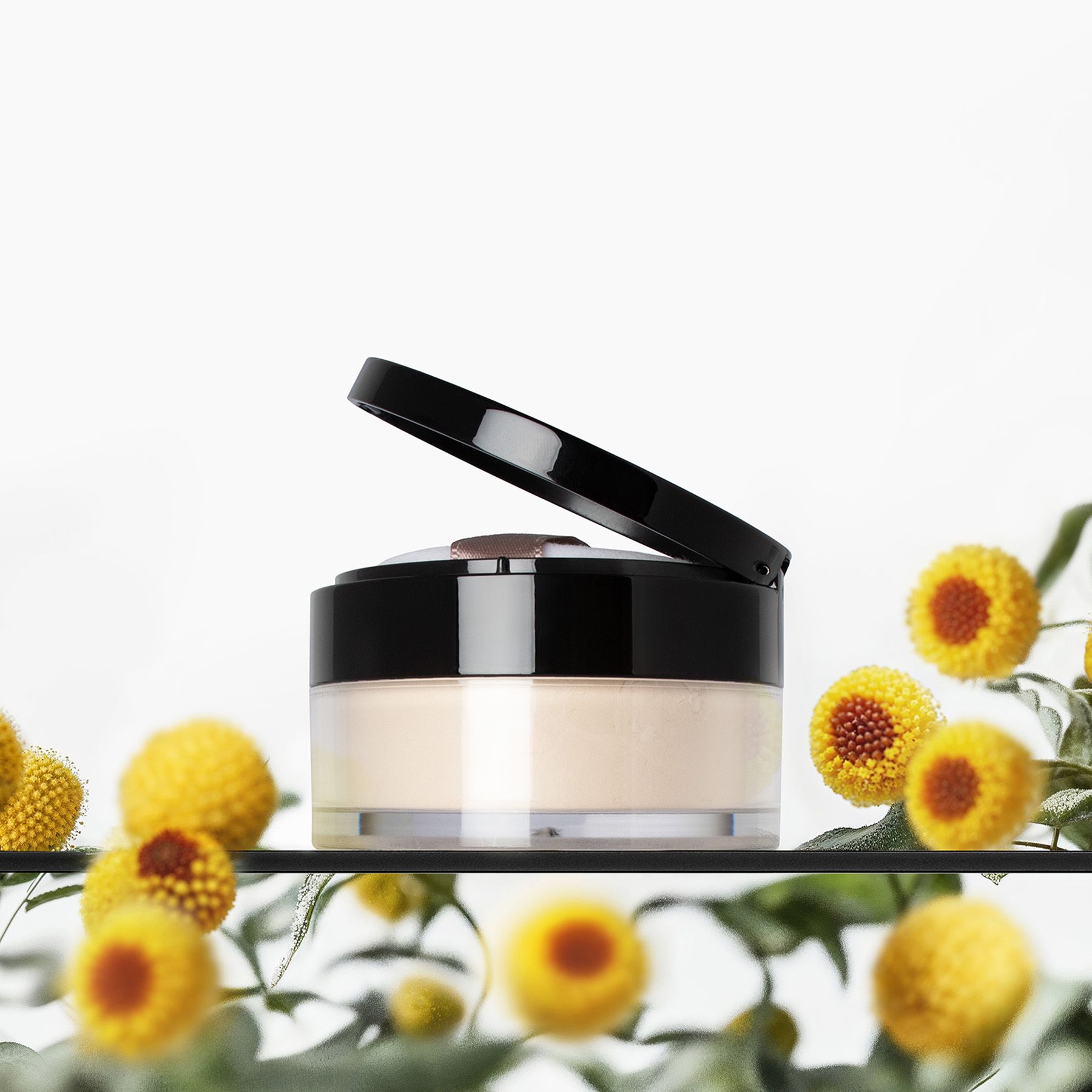 Puder-Dose mit schwarzem Deckel. Gelbe Blumen und grüne Blätter im Hintergrund.