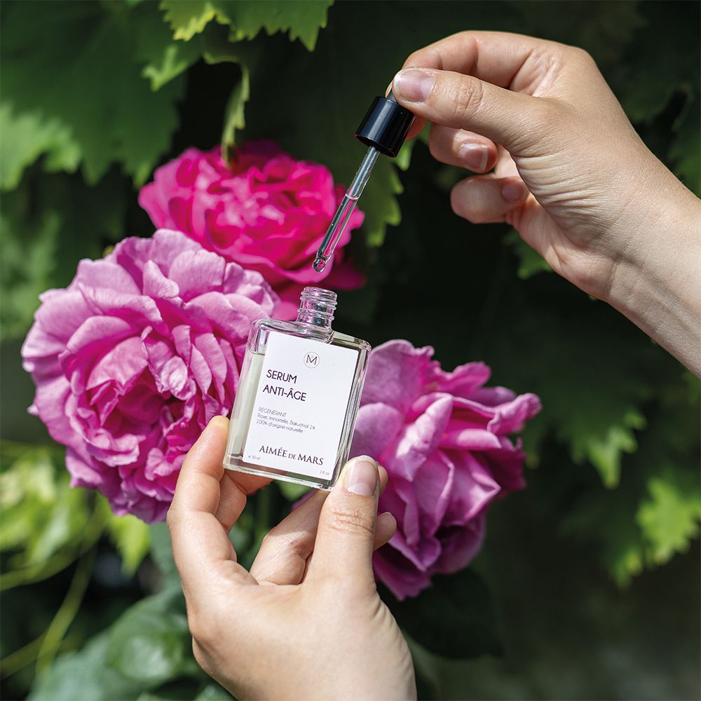 Hände halten Flakon und Pipette über Rosen. Flakon mit "SERUM ANTI-ÂGE" und "AIMÉE DE MARS".