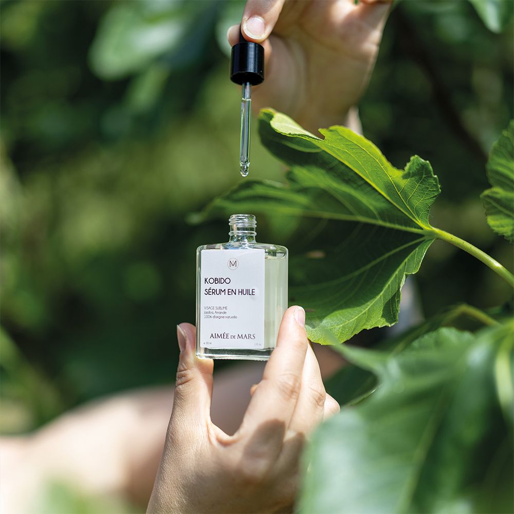 Mains tenant un flacon de sérum huileux et un compte-gouttes au-dessus de feuilles vertes. Gouttes tombant.