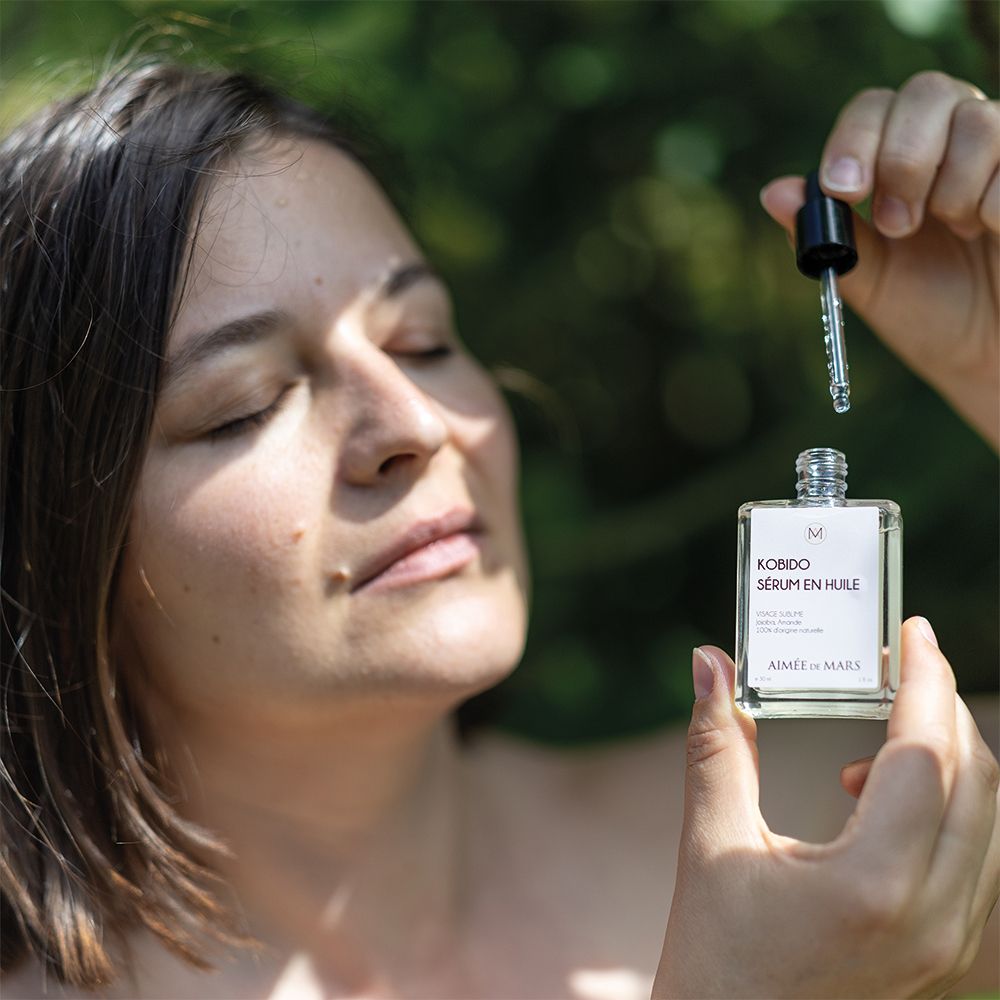 Femme tenant un flacon de sérum huileux et un compte-gouttes. Visage détendu, yeux fermés. Fond vert.