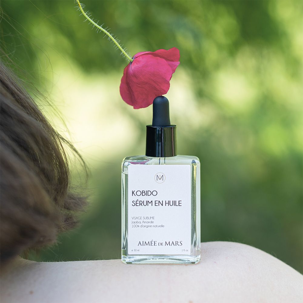 Flacon de sérum huileux et compte-gouttes, placé à côté d'une fleur rouge. Épaule d'une personne en arrière-plan.