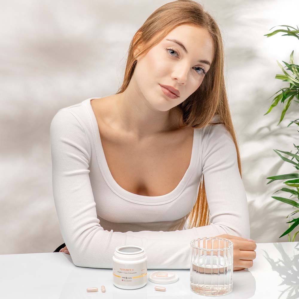 Frau mit Vitamin C-Behälter und Wasserglas. Tabletten liegen daneben. Produktname und Logo Swissells sichtbar.