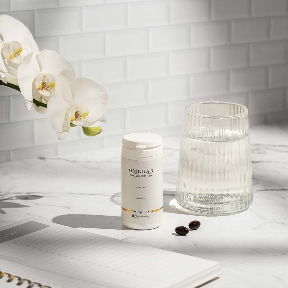 Weiße Dose, Glas Wasser, Kapseln auf Tisch. Dose: OMEGA 3. Hintergrund: Orchideen, Notizbuch.