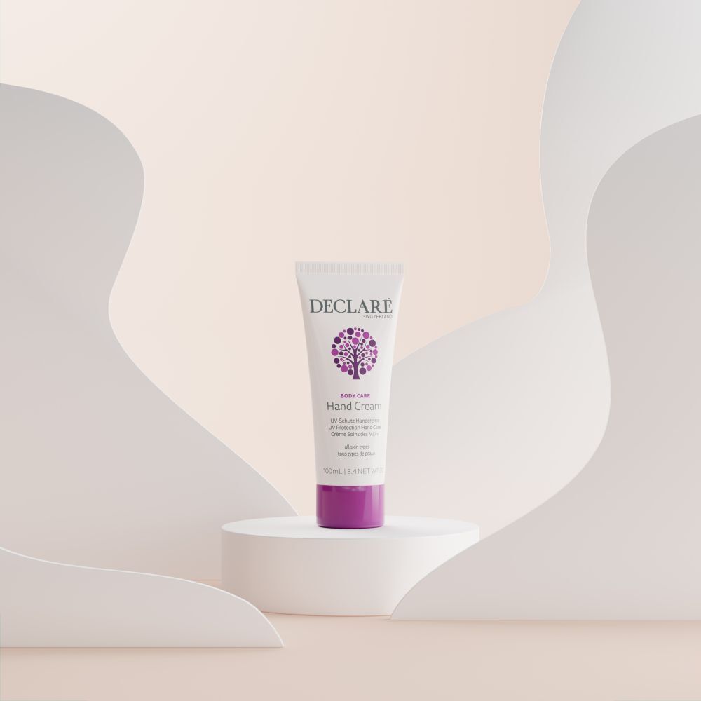 Eine Tube Handcreme auf einem Podest. Weiß, mit lila Akzenten und Logo. Text: DECLARE, Hand Cream, UV Protection, für alle Hauttypen.