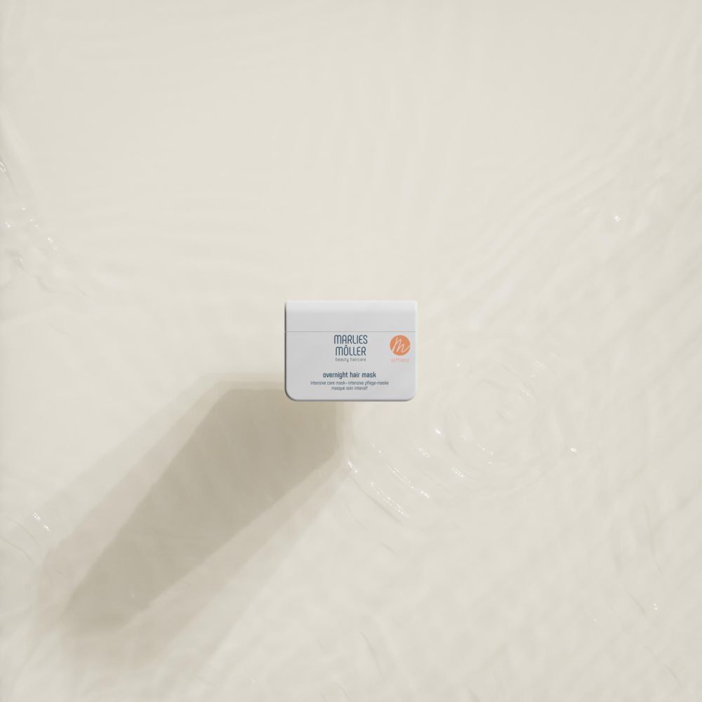Weißer Tiegel mit Aufschrift MARLIES MÖLLER. Text: overnight hair mask. Orangefarbenes Logo. Der Tiegel liegt in Wasser.