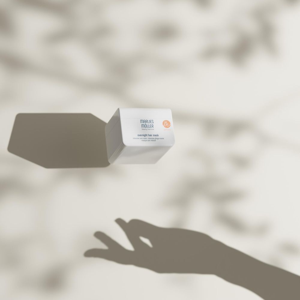 Weißer Tiegel mit Aufschrift MARLIES MÖLLER. Text: overnight hair mask. Orangefarbenes Logo. Der Tiegel schwebt vor einem Schatten.