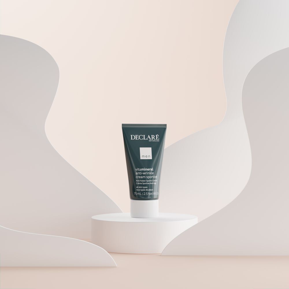 Un tube de DECLARE Men Anti-Wrinkle Cream Sportive sur un socle blanc. Fond: formes abstraites beiges et blanches.