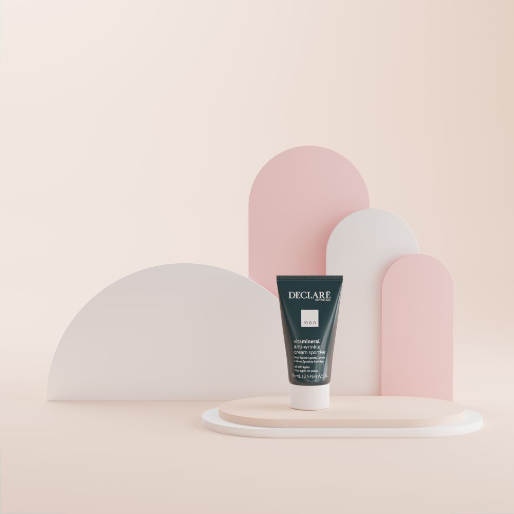 Un tube de DECLARE Men Anti-Wrinkle Cream Sportive sur un socle. Fond: formes abstraites roses, beiges et blanches.
