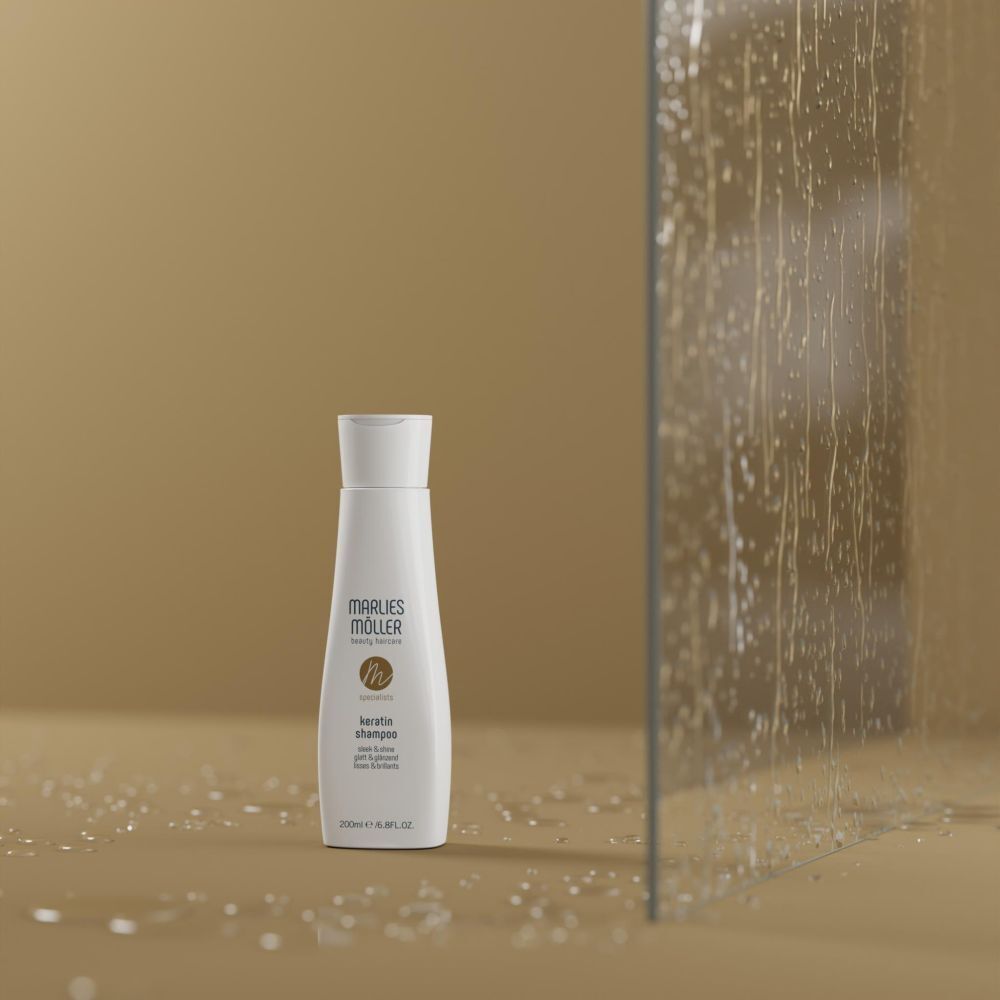 Eine weiße Flasche Marlies Möller Keratin Shampoo Sleek & Shine steht auf einem beigefarbenen Untergrund neben einer nassen Glaswand.