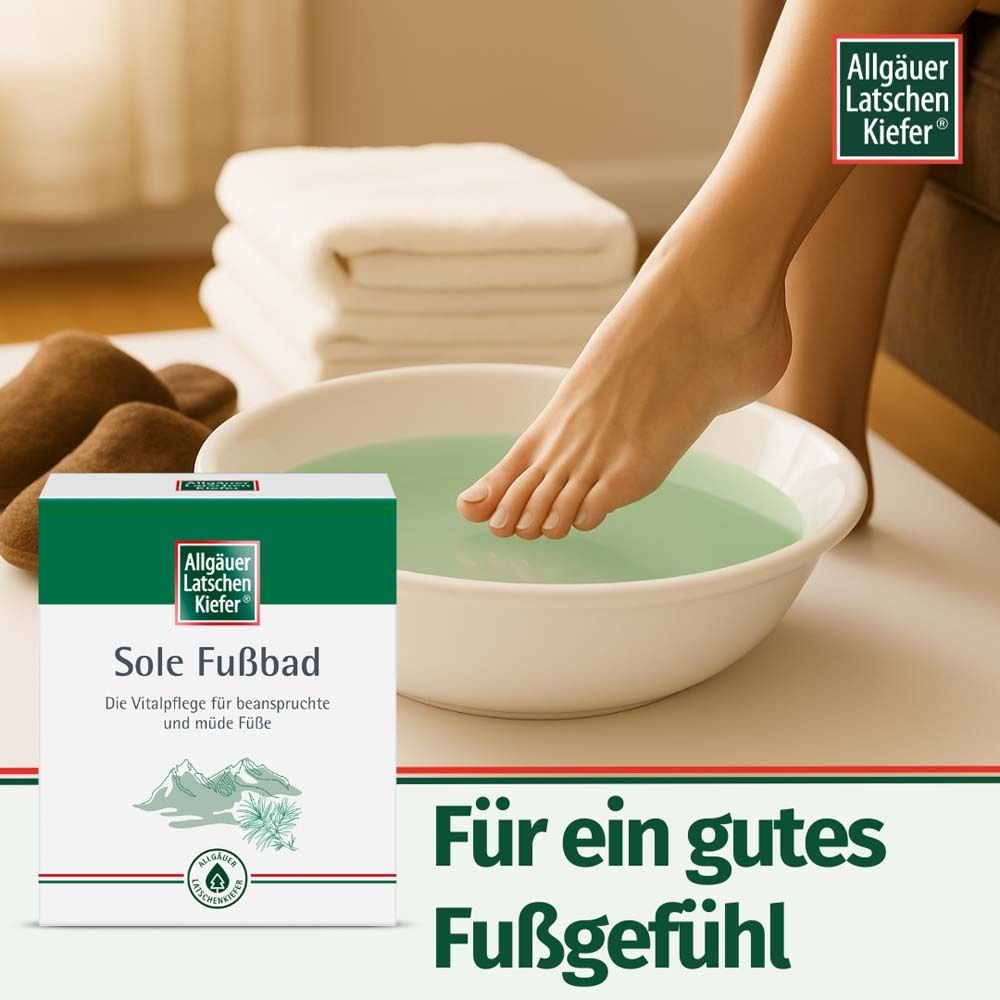 Fuß in einer Schüssel mit Fußbad. Verpackung von Allgäuer Latschenkiefer Sole Fußbad daneben. Handtücher und Hausschuhe.
