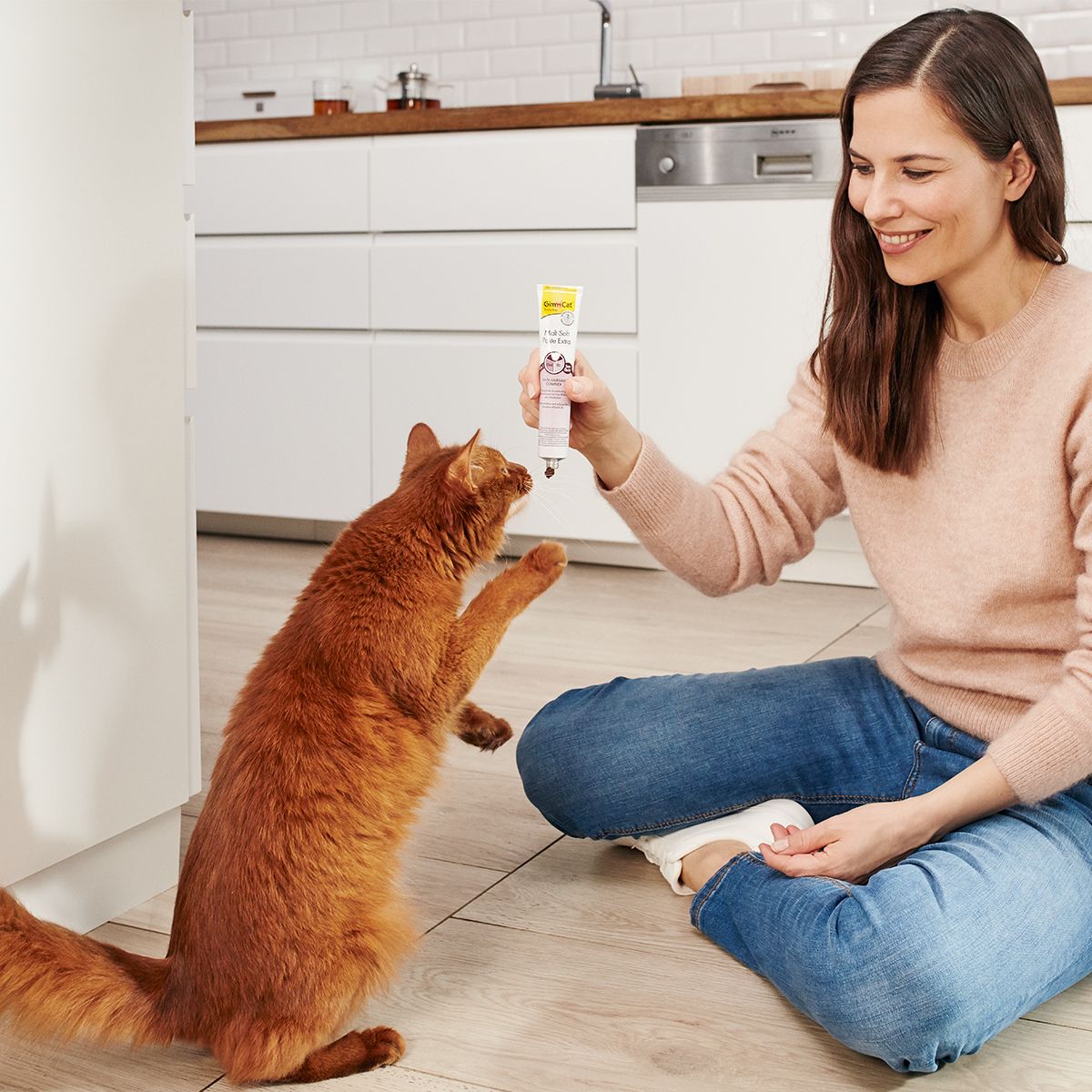 Femme et chat dans une cuisine. La femme tient un tube de GimCat Malt-Soft Paste Extra. Le chat saute. Rose, jaune et blanc.