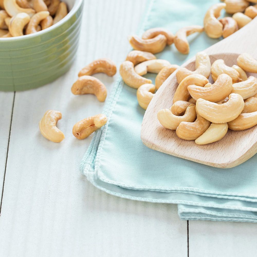 Noix de cajou sur une cuillère en bois et sur un tissu bleu. Bol vert en arrière-plan.