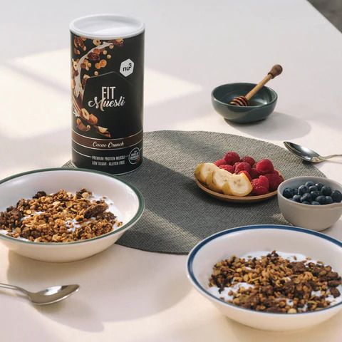 nu3 Fit Müsli Cacao Crunch Dose und Schalen mit Müsli, Obst. Auf Tisch. Blaubeeren, Himbeeren, Banane, Honig.