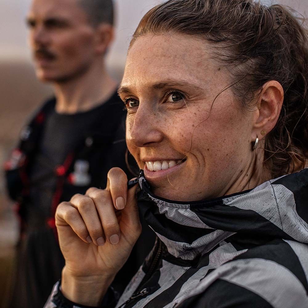 Porträt einer lächelnden Frau in Sportkleidung. Im Hintergrund eine weitere Person. Sonnenlicht.