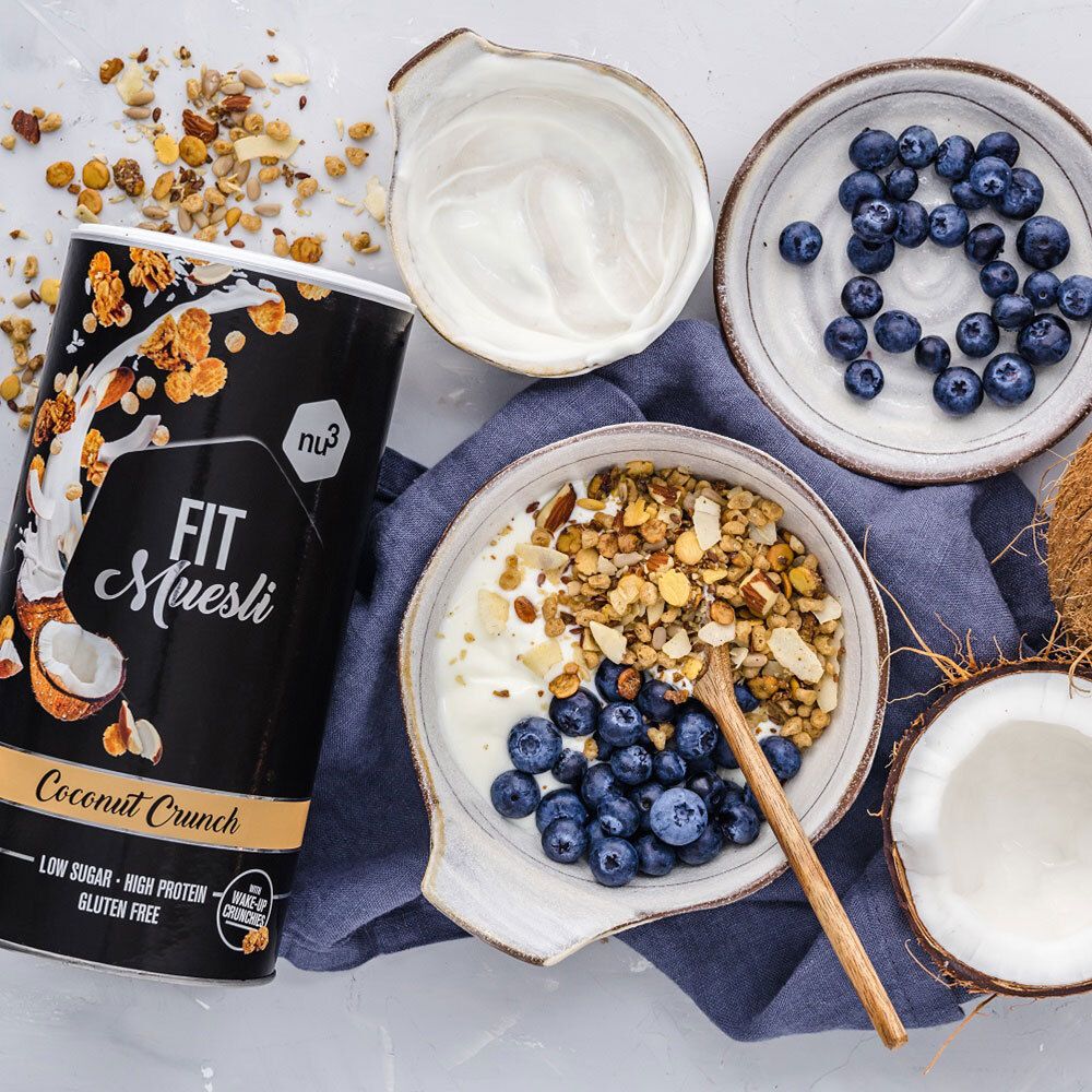 Boîte noire nu3 Fit Müsli, Coconut Crunch, à côté d'un bol de muesli, myrtilles, yaourt. Noix de coco et cuillère en bois sur la table.