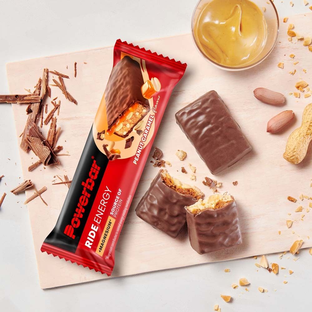 Barre PowerBar Ride Energy, Peanut Caramel. Sur planche de bois. Barre ouverte, cacahuètes et beurre de cacahuète en arrière-plan.