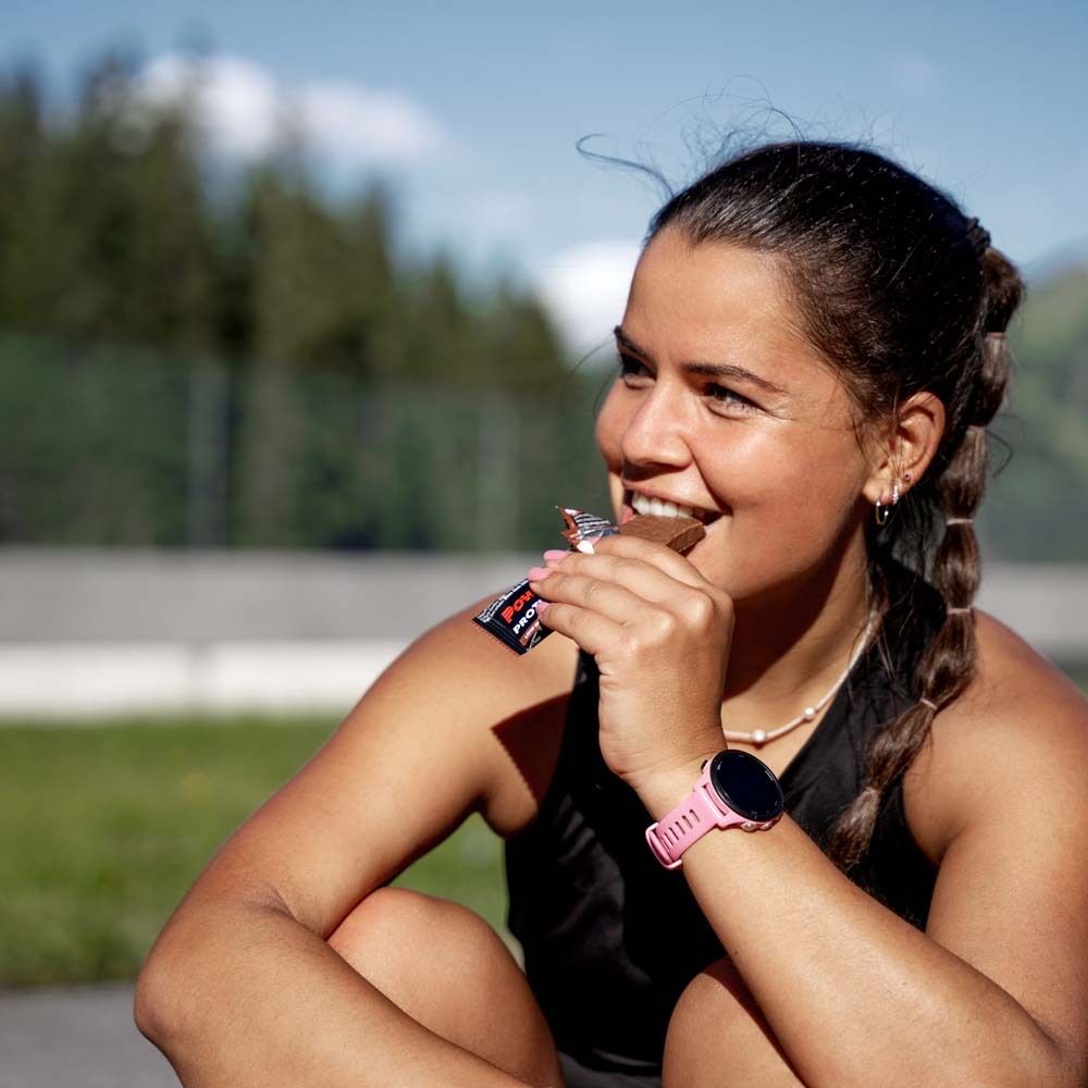 Une femme mange une barre Powerbar Protein+. Elle est assise à l'extérieur. Elle porte une montre intelligente.
