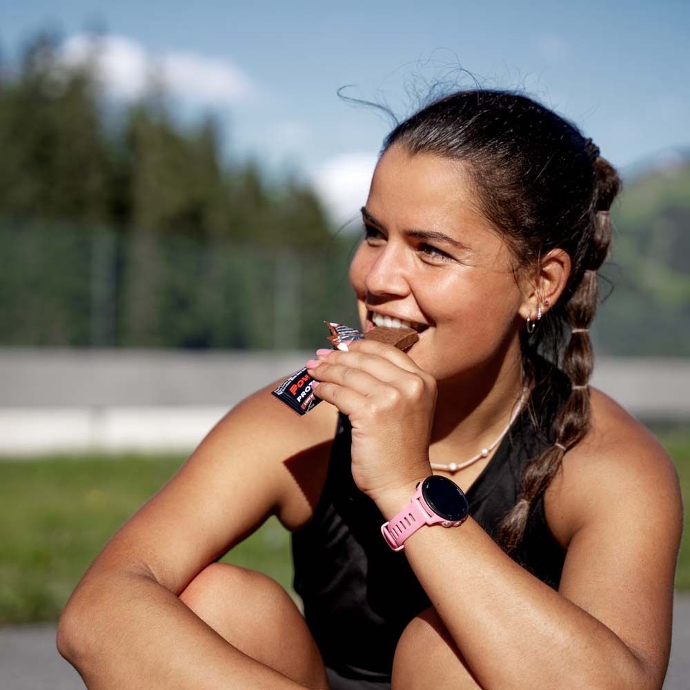 Femme mange une barre Powerbar Protein+ Fibre. Emballage rose. Elle porte une montre connectée. En extérieur.