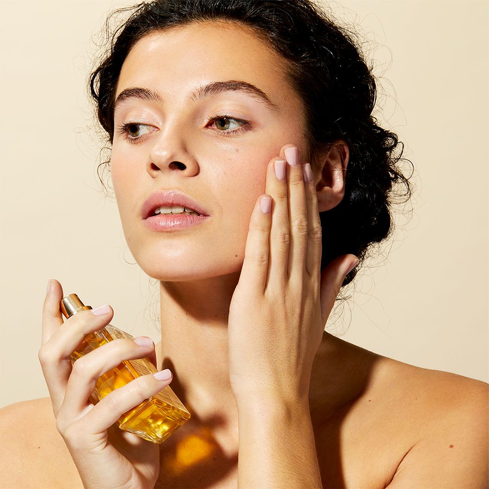 Femme appliquant de l'huile sur le visage. Flacon NUXE Huile Prodigieuse® visible. Peau claire.