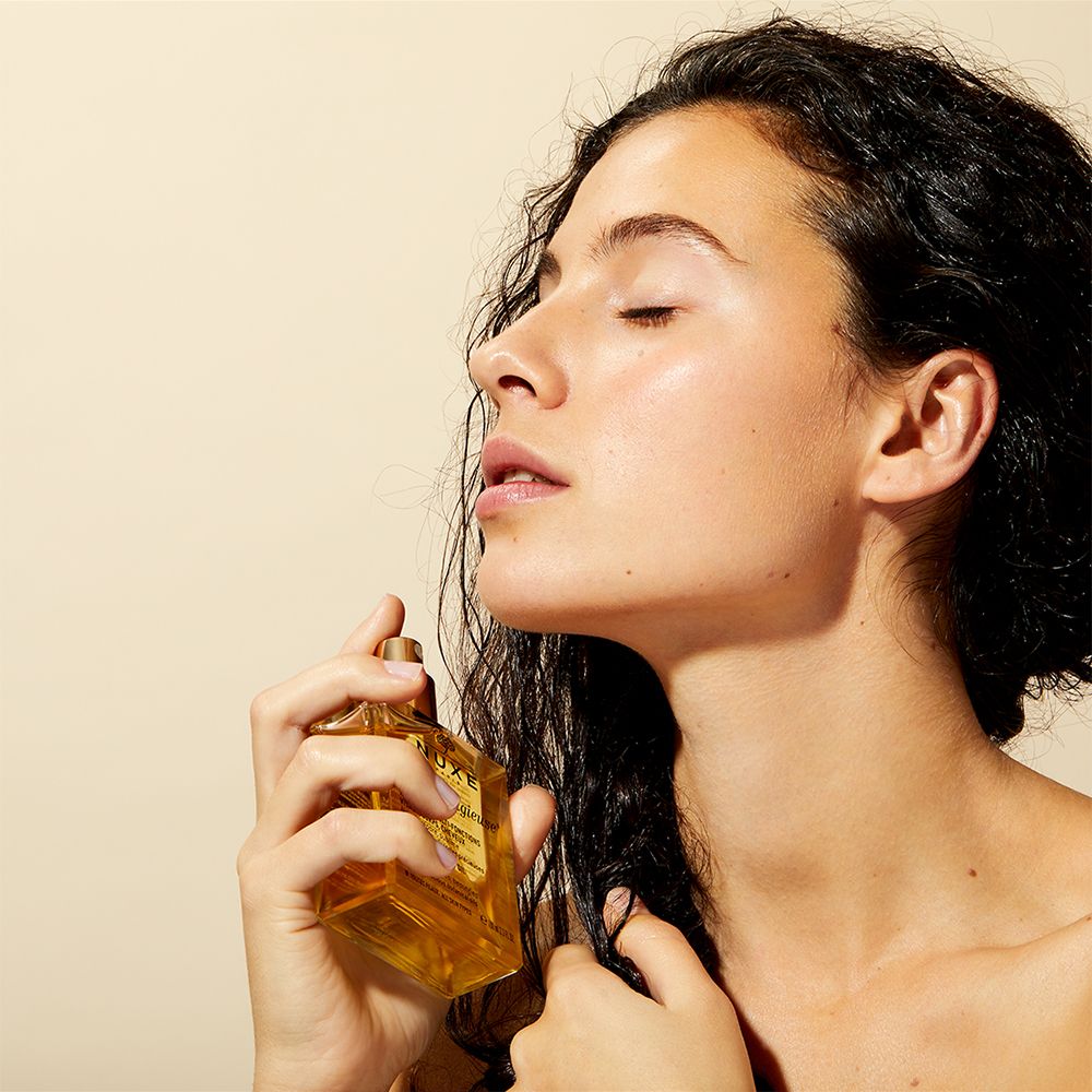 Femme appliquant de l'huile sur les cheveux. Flacon NUXE Huile Prodigieuse® visible. Cheveux foncés.