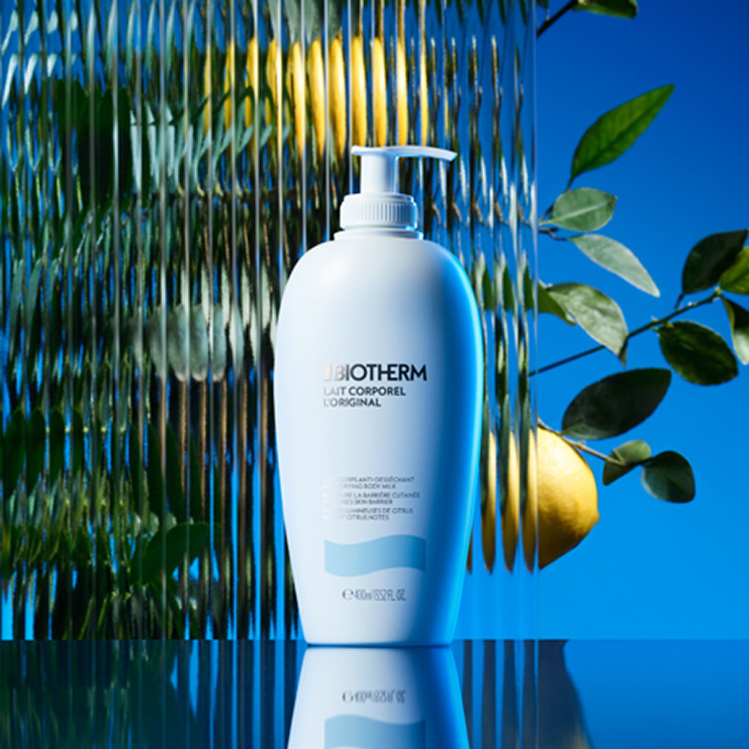 Eine weiße Flasche mit Pumpverschluss vor blauem Hintergrund. Text: BIOTHERM Lait Corporel L'Original. Gelbe Zitrone.