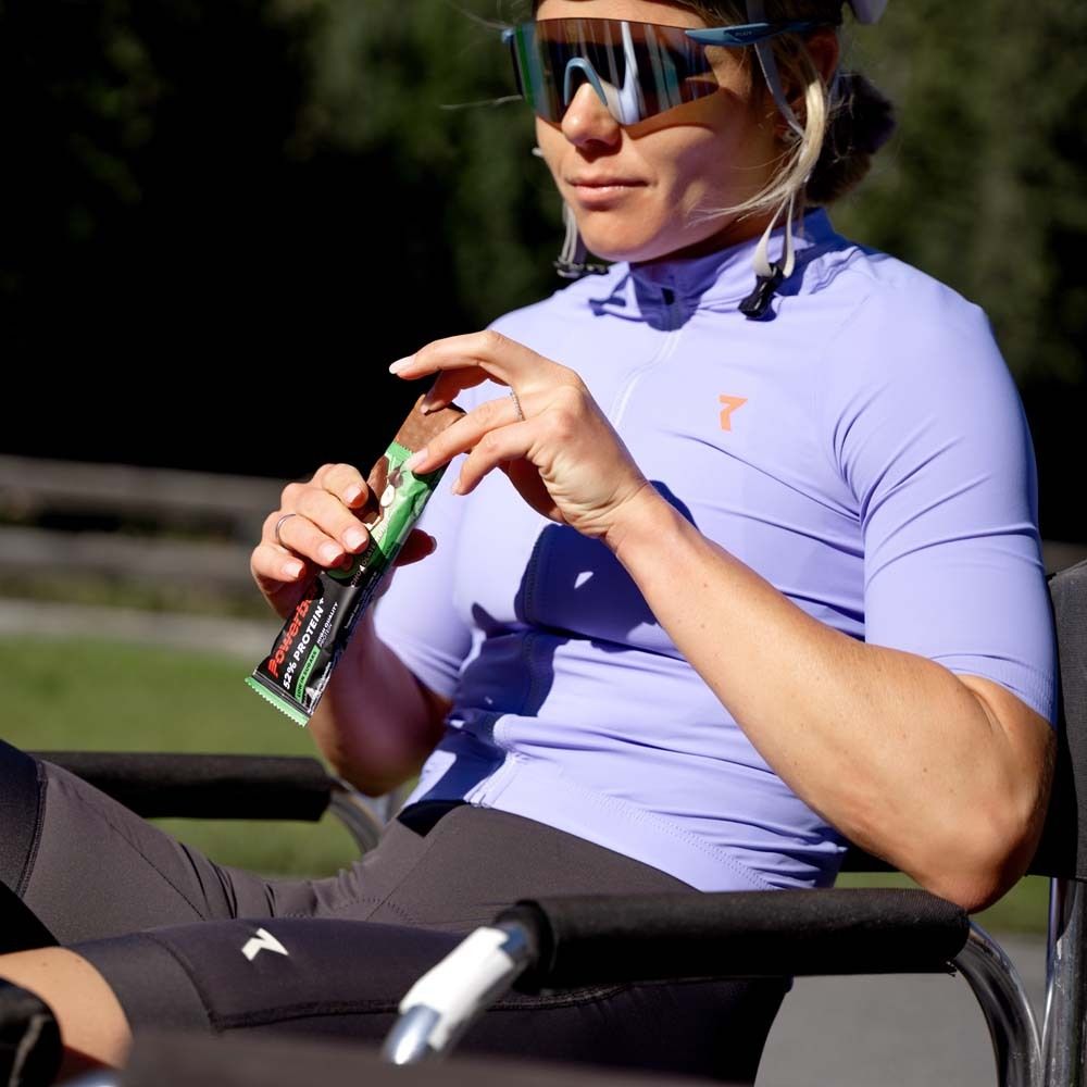 Person hält eine grüne Powerbar 52% Protein Plus Riegel. Person trägt Sportkleidung und Sonnenbrille.