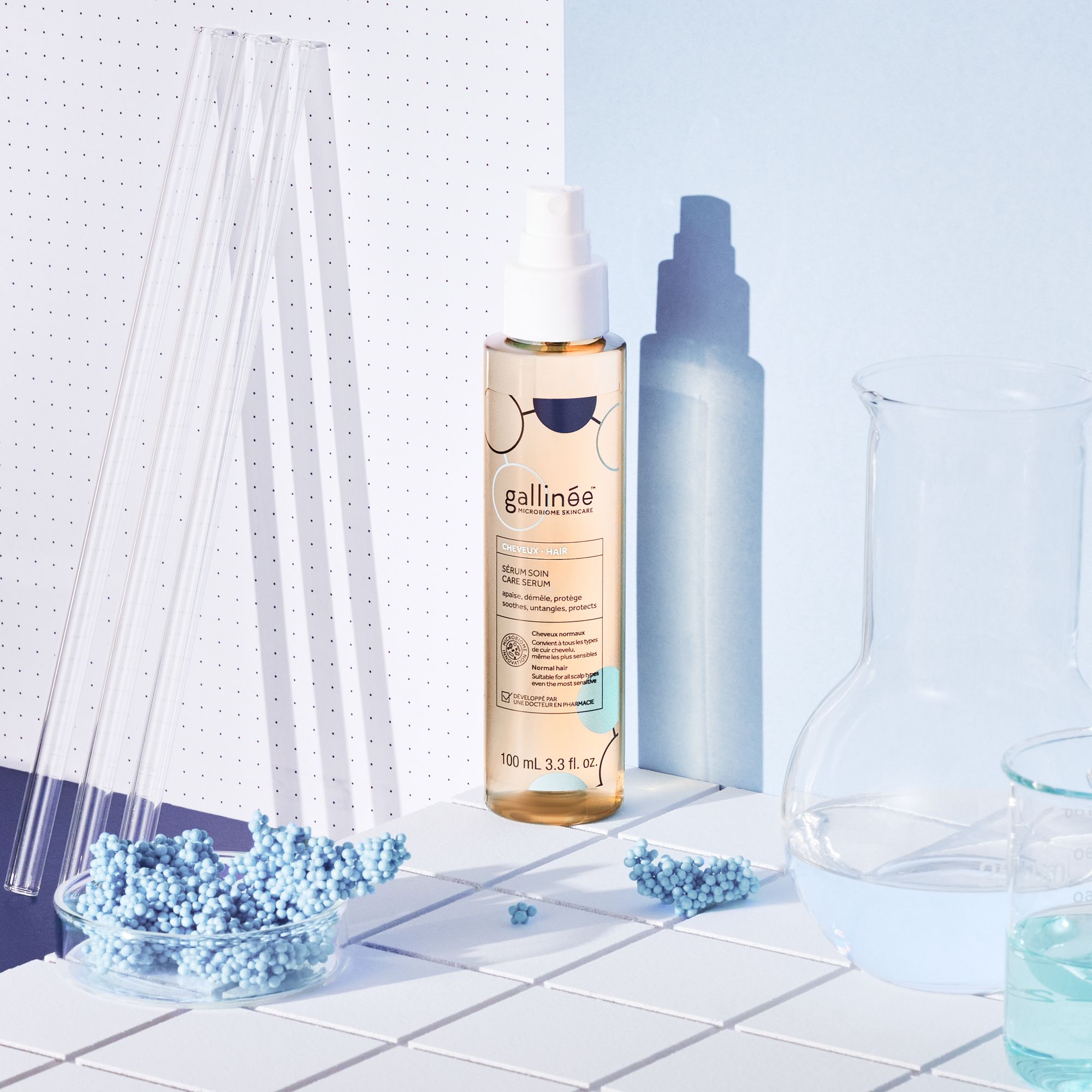 Flacon de sérum capillaire avec vaporisateur. Sur fond bleu et blanc avec matériel de laboratoire.