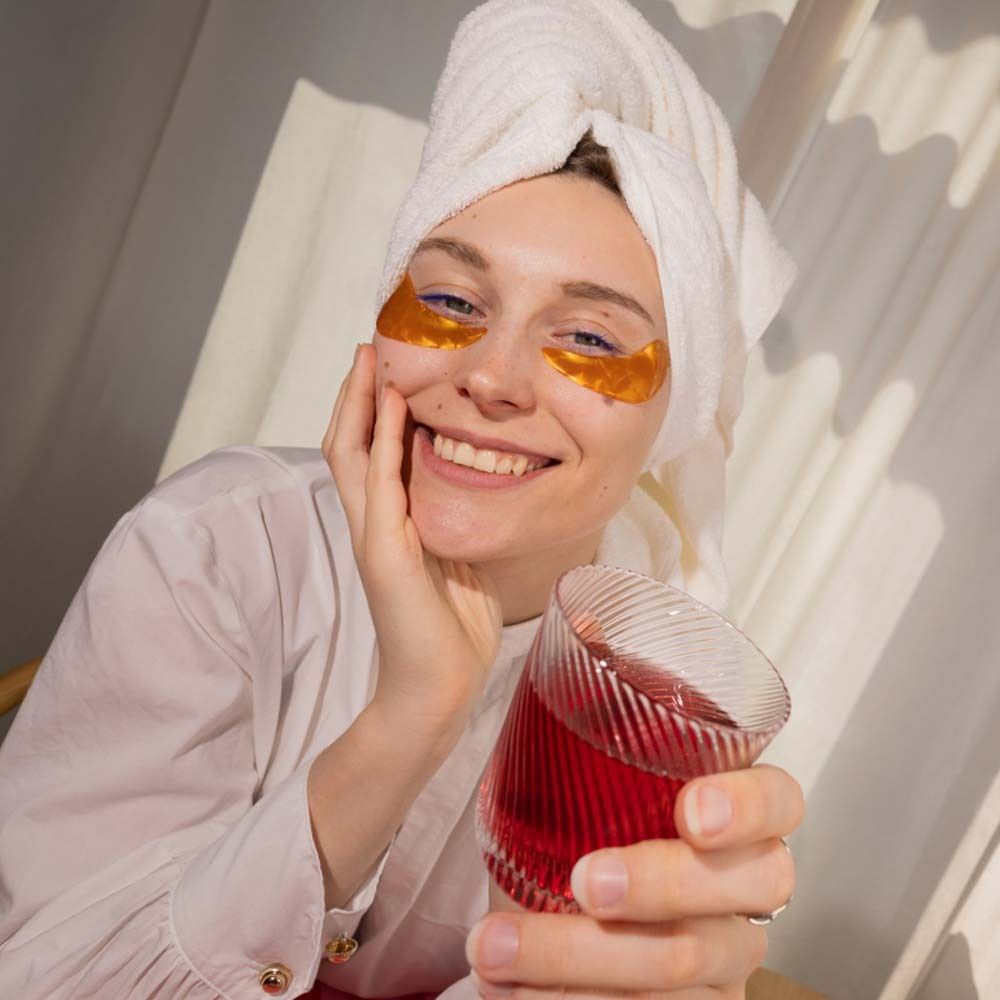 Frau mit Augenpads hält Glas mit rotem Getränk. Im Hintergrund eine Verpackung.