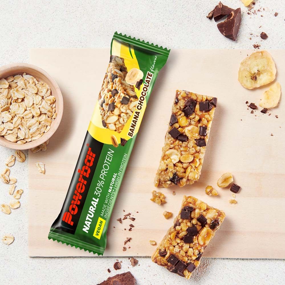 Powerbar NATURAL PROTEIN Bananen-Schokolade Riegel auf Holzbrett mit Haferflocken und Bananenscheiben.