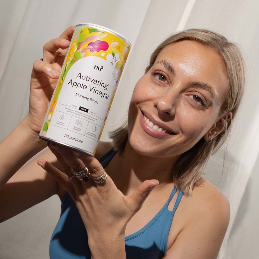 Femme tenant une boîte "nu3 Activating Apple Vinegar". Nom du produit et logo visibles. La femme sourit.