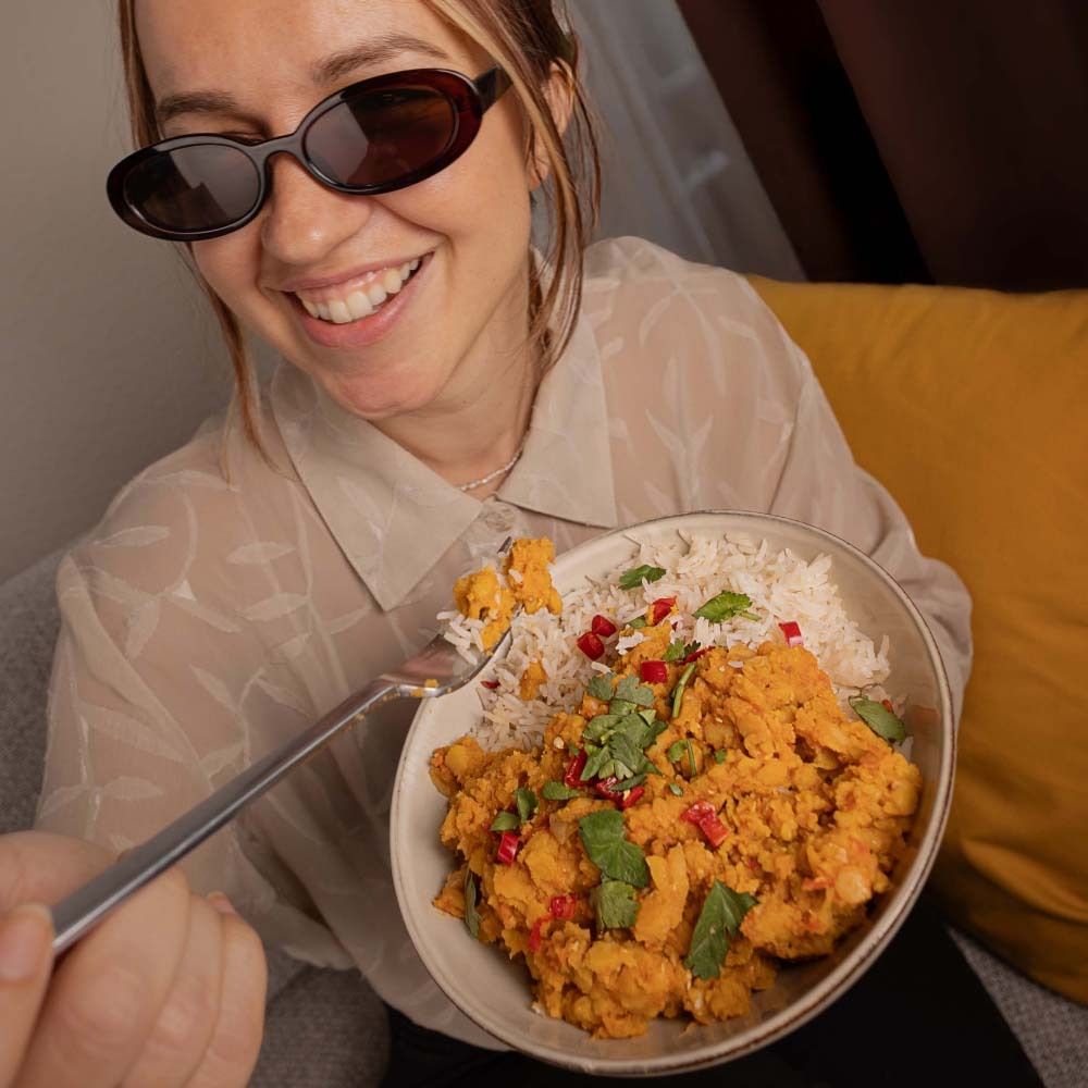 Person hält eine Schüssel mit Reis und Curry. Frau mit Sonnenbrille isst Curry. Sichtbar: Curry, Reis, Koriander.