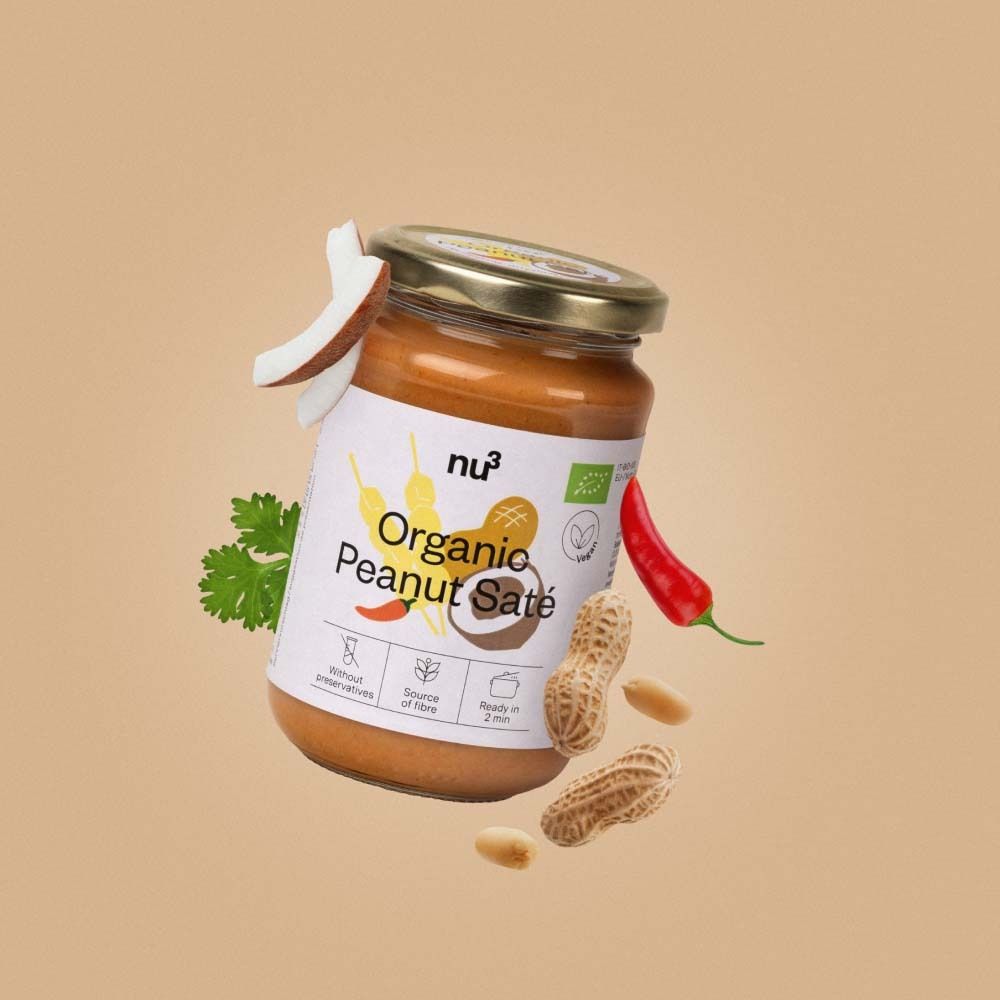 Glas mit Erdnuss-Saté-Sauce, umgeben von Erdnüssen, Kokosnuss-Stücken, Chili und Koriander. Aufschrift: Organic Peanut Saté.