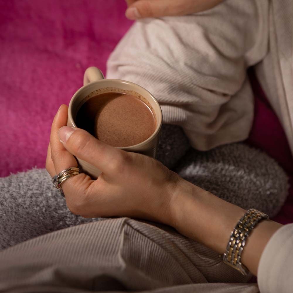 Mains tenant une tasse avec une boisson. Boîte "Balancing Hot Cocoa" non visible. Personne assise sur une couverture rose.