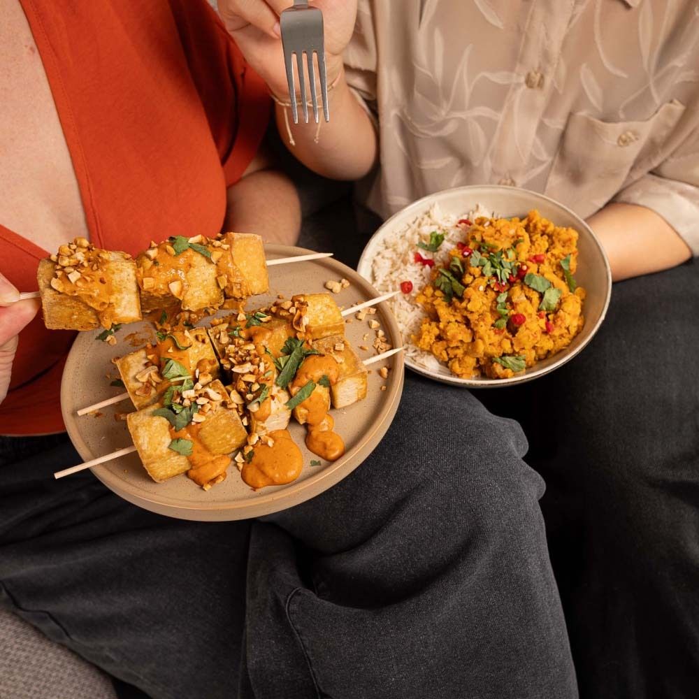 Personnes mangeant des brochettes de tofu à la sauce saté aux cacahuètes et au curry de lentilles. Brochettes sur une assiette, curry dans un bol.