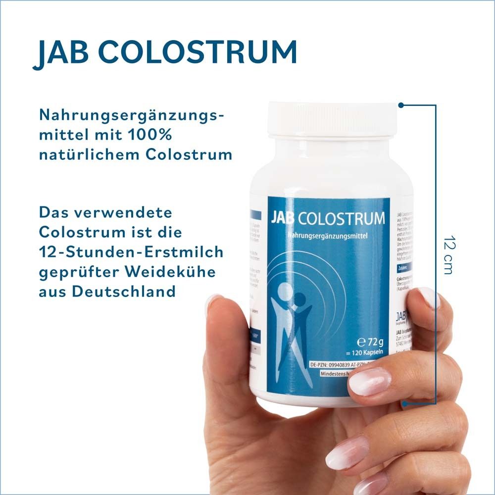Main tenant un flacon blanc de gélules. Inscription: JAB Colostrum. 12 cm de haut.