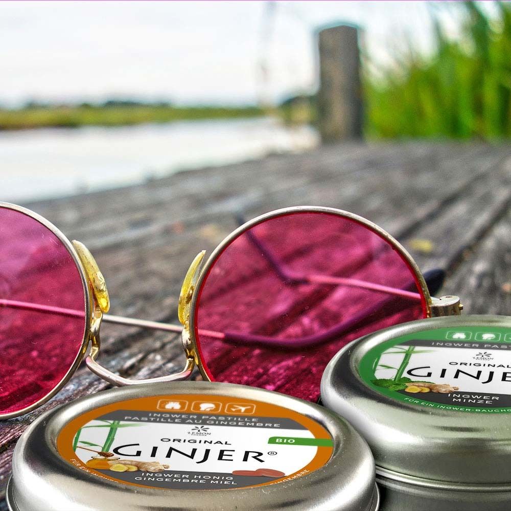 Zwei Dosen Ingwer-Pastillen neben einer Sonnenbrille. Etiketten mit Text und Logos. Eine Dose mit "BIO"-Siegel.