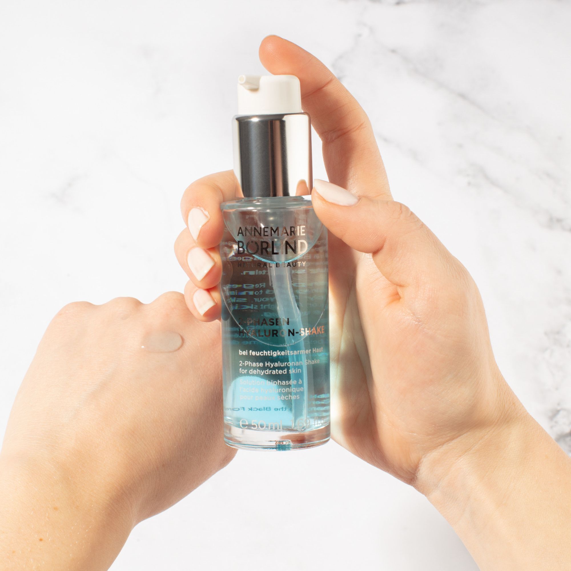 Hände halten ANNEMARIE BÖRLIND 2-Phasen Hyaluron-Shake. Tropfen auf Hand. Pumpflasche.