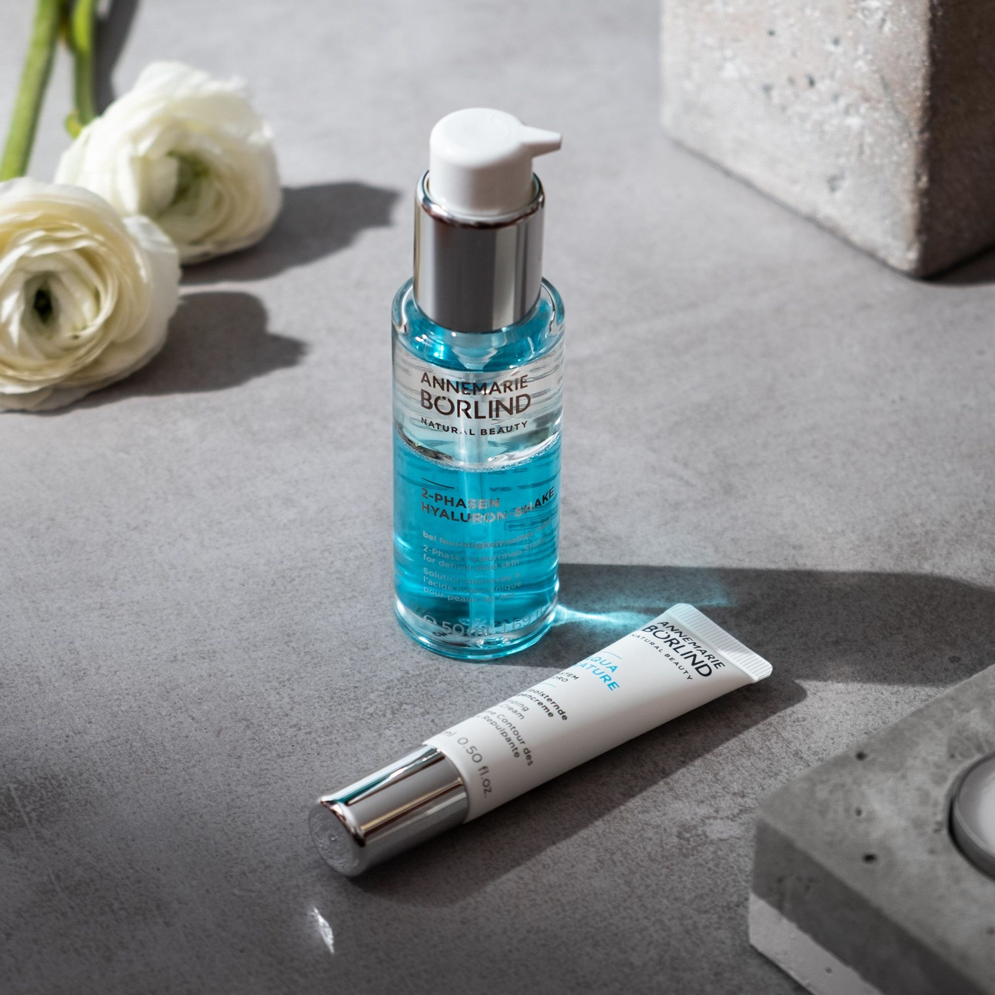 ANNEMARIE BÖRLIND 2-Phasen Hyaluron-Shake und Tube. Auf grauer Oberfläche. Weiße Blüten.