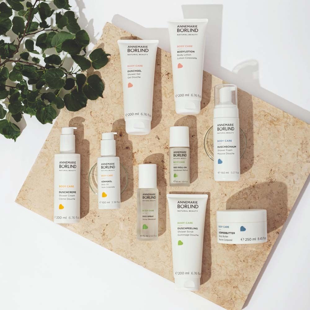 Verschiedene weiße Flaschen und Tiegel. Aufschrift: ANNEMARIE BÖRLIND, BODY CARE. Gelbes Logo. Auf Steinplatte.