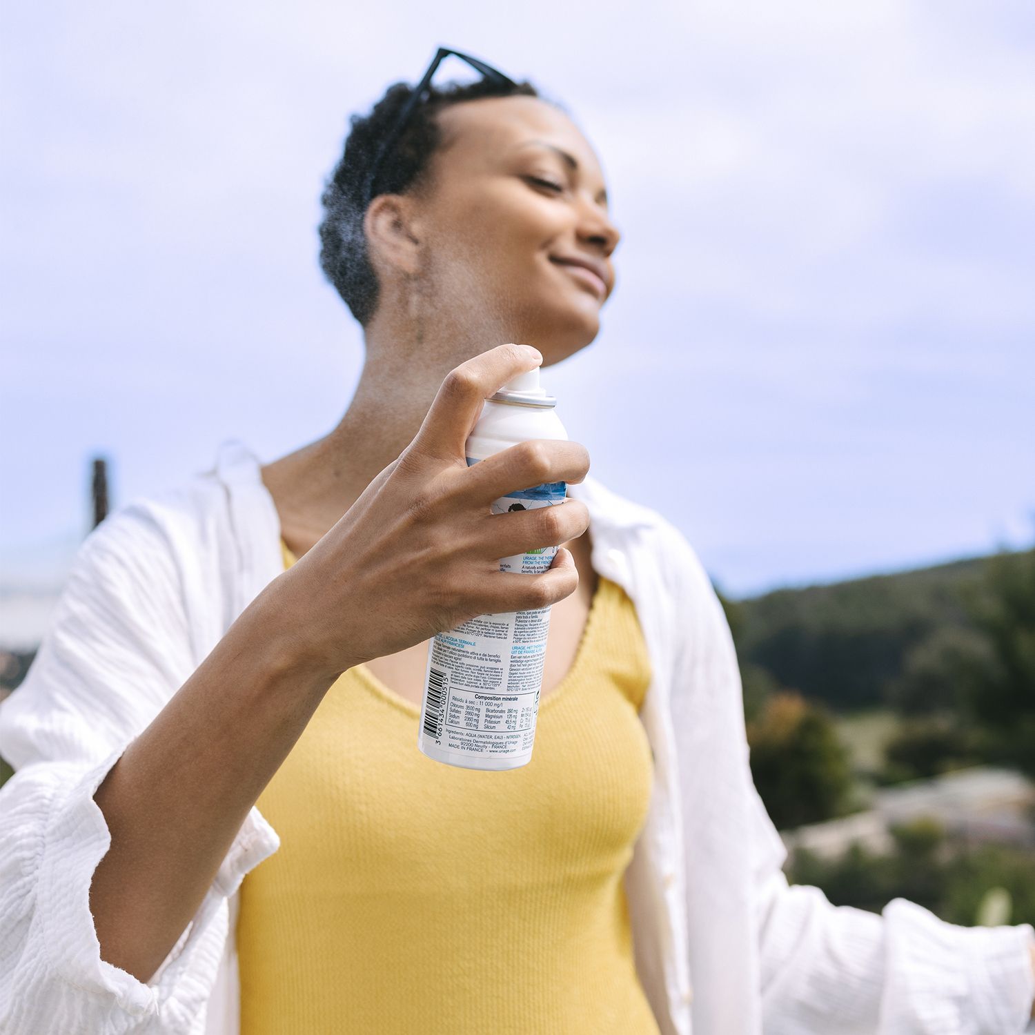 Frau sprüht Uriage Eau Thermale auf ihr Gesicht. Weiße Flasche, blauer Deckel. Hintergrund: Natur.