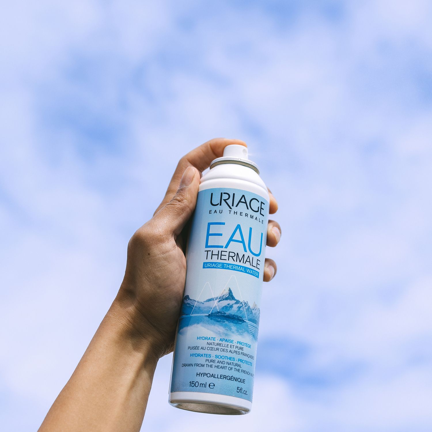 Hand hält Uriage Eau Thermale Spray. Blaue und weiße Flasche. Hintergrund: blauer Himmel.