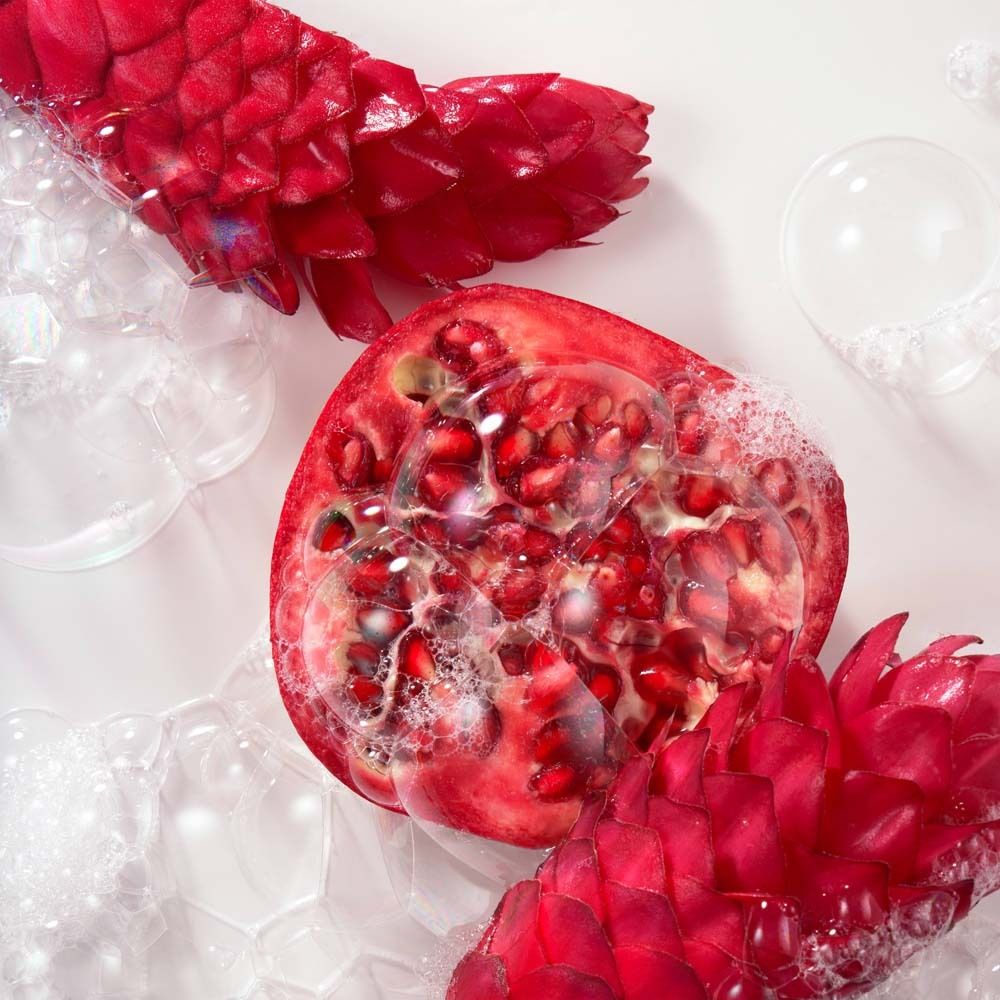 Nahaufnahme einer halbierten Granatapfelfrucht mit Kernen, umgeben von Seifenblasen und roten Blüten.
