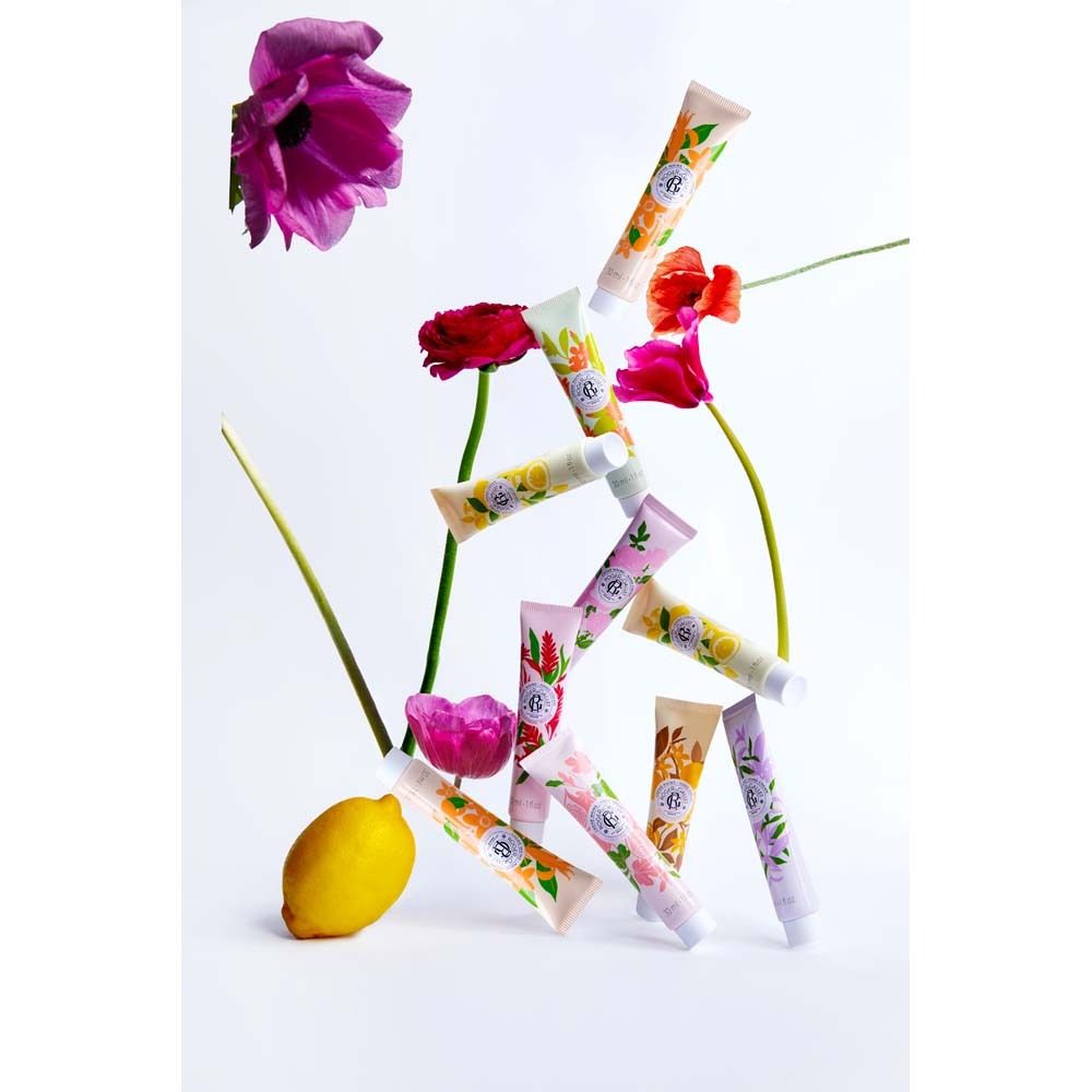 Plusieurs tubes de crème pour les mains, couleurs et motifs variés, empilés. Quelques fleurs et un citron à côté. Différentes marques.